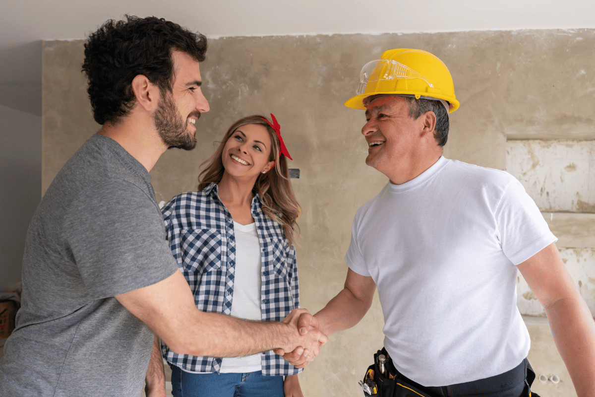 general contractor in a white t-shirt and yellow helmet shaking hands with a couple